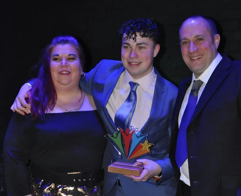 Three people pose together at an awards ceremony, smiling towards the camera against a dark stage backdrop lit with blue and purple lighting. The person in the centre wears a light blue suit and tie and holds a colourful trophy featuring stylised figures and a star. The two people beside them are dressed in formal evening attire, with one person’s arm around the award winner. The image captures a celebratory moment following a presentation or recognition event.
