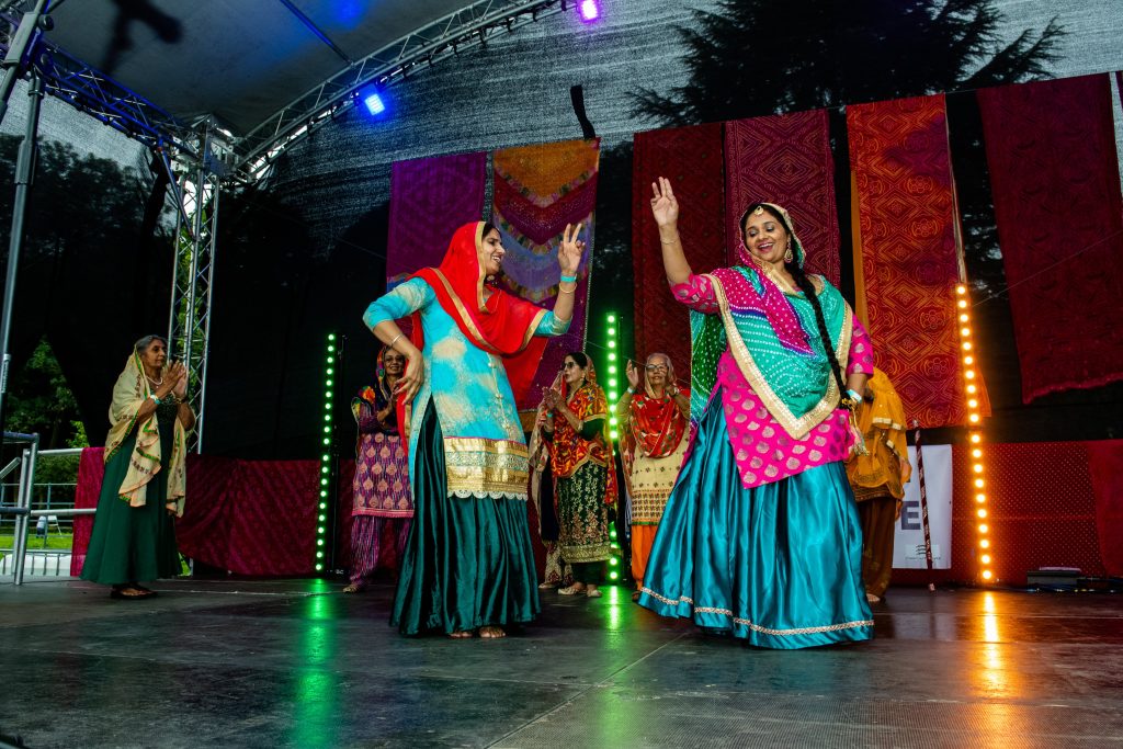 A group of performers in brightly coloured traditional Punjabi clothing dance on an outdoor stage during the Festival of Thetford & Punjab. Two dancers in turquoise, pink and red outfits perform at the front while others clap and dance behind them. Colourful textile panels hang as a backdrop, with stage lighting illuminating the performance.