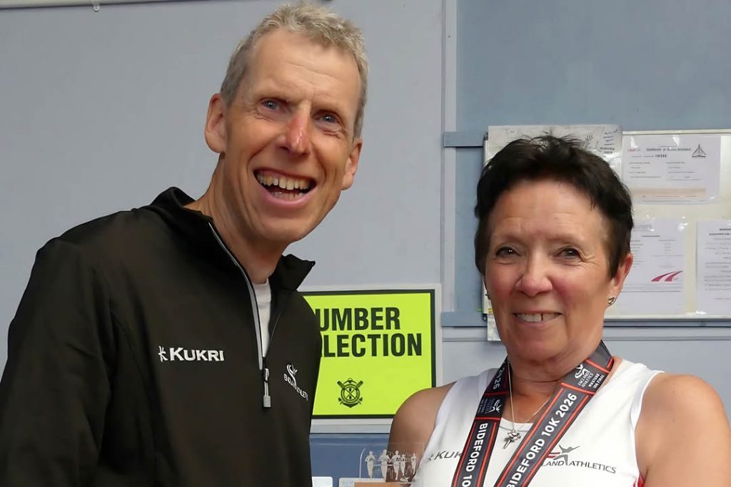 A smiling man in a black Kukri sports jacket stands beside a woman wearing a white athletics vest and a medal ribbon around her neck. They are indoors in what appears to be a sports clubhouse or event venue, with notices and a bright yellow “Number Collection” sign visible in the background. The woman is holding a small trophy or award.