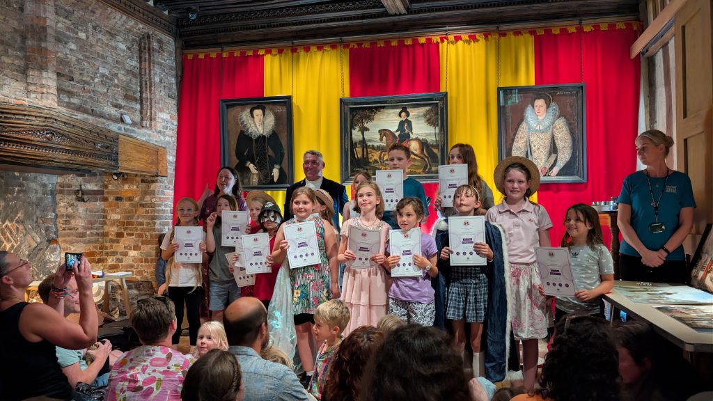 A group of children proudly hold certificates during an event at Ancient House Museum, standing in front of historic portraits and red and yellow drapes.