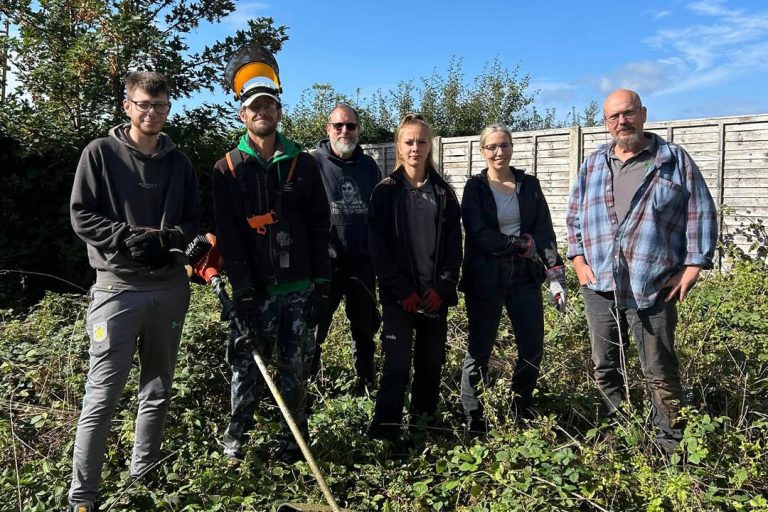 Six volunteers stand in an overgrown outdoor area holding gardening and clearing tools during a community clean-up session. One person is wearing protective headgear and carrying a strimmer, while others wear gloves and outdoor work clothes. A wooden fence and trees can be seen behind them under a bright blue sky.