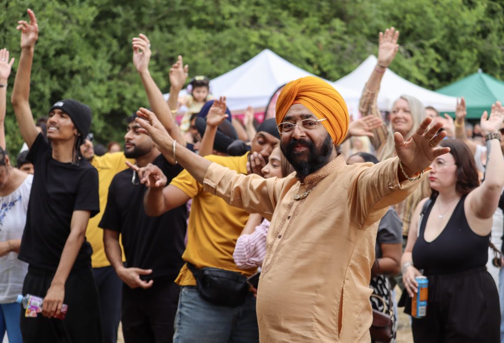 A lively crowd dances outdoors at the Festival of Thetford & Punjab, with a man in an orange turban and traditional dress dancing in the foreground.