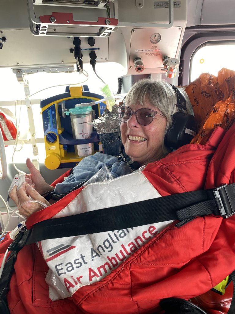 A smiling woman sits strapped into a medical stretcher inside an East Anglian Air Ambulance helicopter. Wearing a headset and wrapped in a red insulated cover, she gives a thumbs-up while surrounded by specialist medical equipment, including monitoring and suction devices mounted inside the cabin.