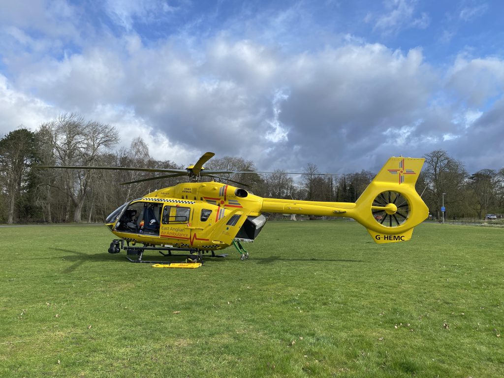 A bright yellow East Anglian Air Ambulance helicopter is parked on a grassy field beneath a dramatic sky of blue patches and dark clouds. The aircraft’s side door is open, revealing the medical cabin, while equipment is laid out on the grass beside it. Trees line the background.