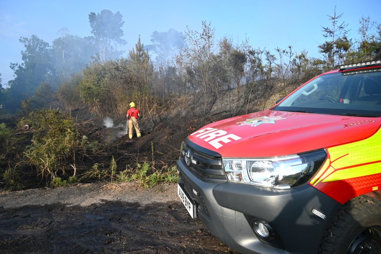 A firefighter in protective clothing tackles smouldering hotspots on a recently burned heathland, while a Norfolk Fire and Rescue Service response vehicle is parked in the foreground. Charred vegetation, smoke and scorched trees stretch across the scene following a wildfire.