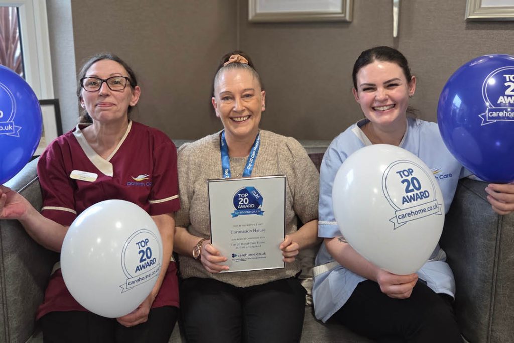 Three members of staff at Care UK’s Coronation House care home in Thetford smile while celebrating the home’s carehome.co.uk Top 20 Award. The woman in the centre holds the award certificate, recognising Coronation House as one of the top 20 care homes in the East of England, while the two women beside her hold blue and white celebratory balloons.