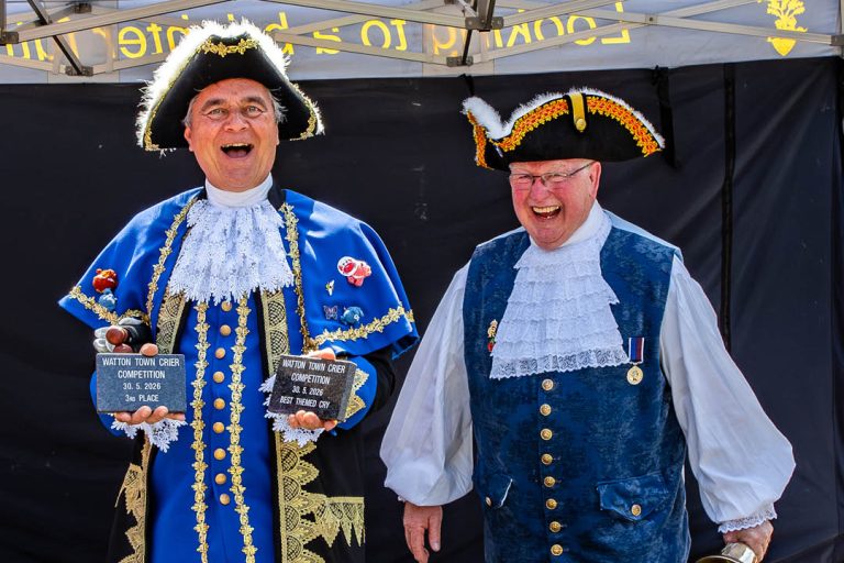 Two smiling town criers in traditional ceremonial dress stand together at the Watton Town Crier Competition. The crier on the left wears an ornate blue and gold uniform and holds awards for third place and Best Themed Cry, while the crier beside him wears a blue waistcoat, white ruffled shirt and black tricorn hat.