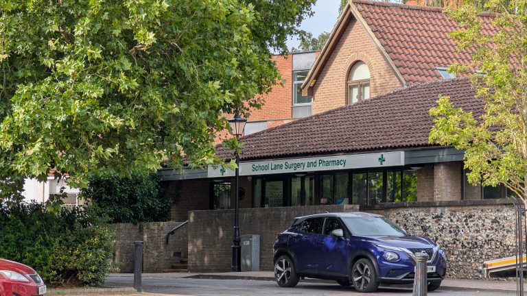 Exterior of School Lane Surgery and Pharmacy in Thetford, with the building partly framed by trees and a car parked outside.