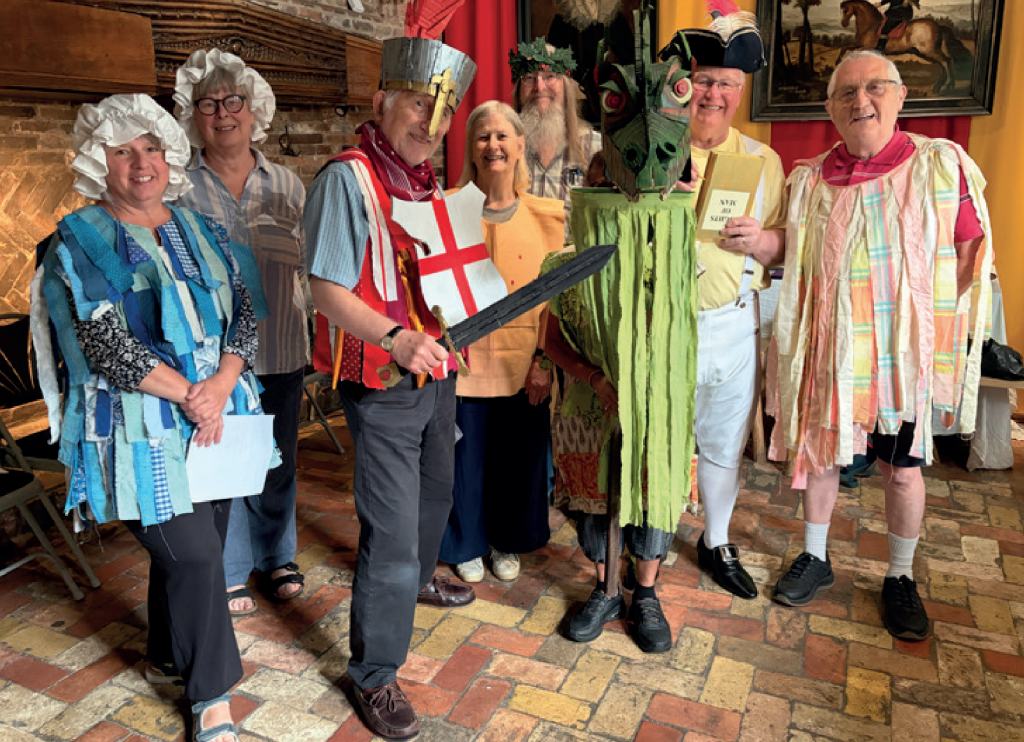 Seven people pose together in colourful costumes inside Ancient House Museum in Thetford, including characters dressed as a knight, a dragon, a historical figure and townspeople.