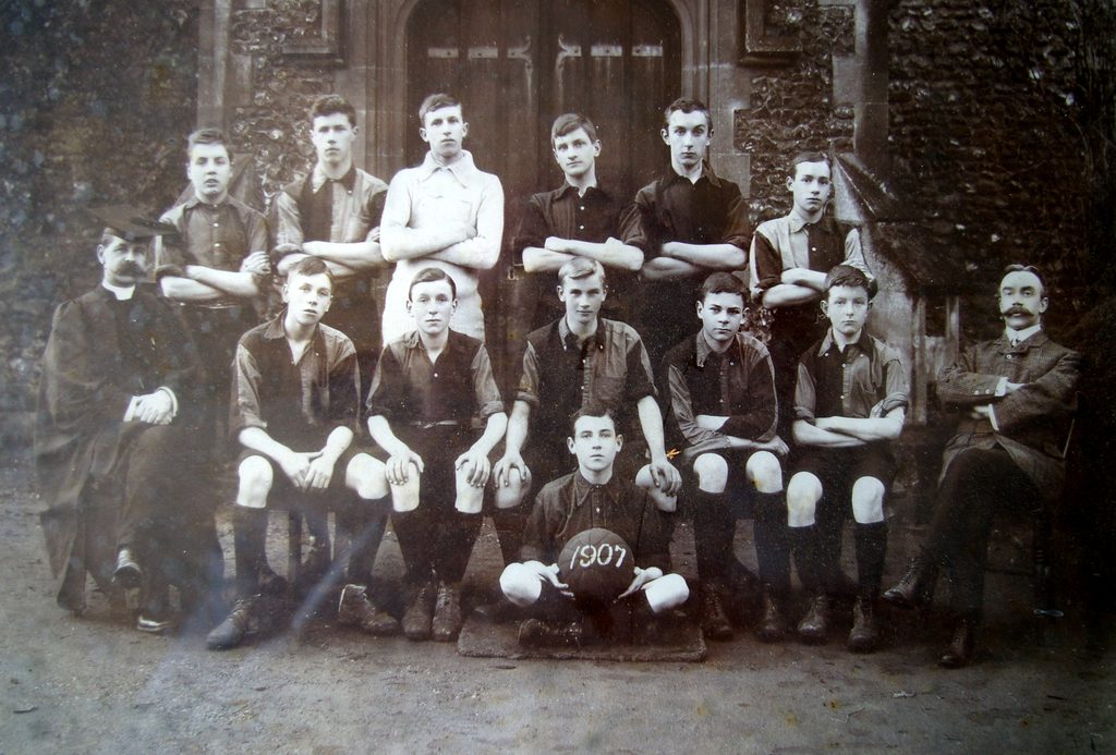 Black-and-white photograph of Thetford Grammar School’s boys’ football team in 1907, with pupils and two members of staff posed outside the school. A pupil seated at the front holds a football marked “1907”.