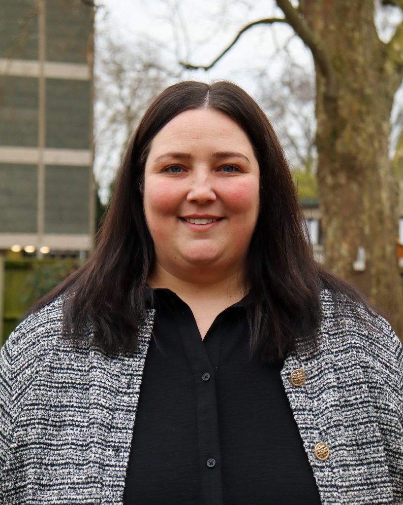 Portrait of a smiling woman with long dark hair, wearing a black blouse and patterned jacket, photographed outdoors.