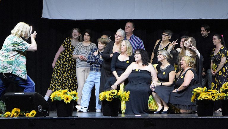 A group of cast members, friends and supporters gather on stage after a performance of Calendar Girls: The Musical at The Carnegie, Thetford, posing for a commemorative photograph. A photographer stands to the left taking the picture, while the group smiles and makes playful poses among pots of bright yellow sunflowers decorating the front of the stage.