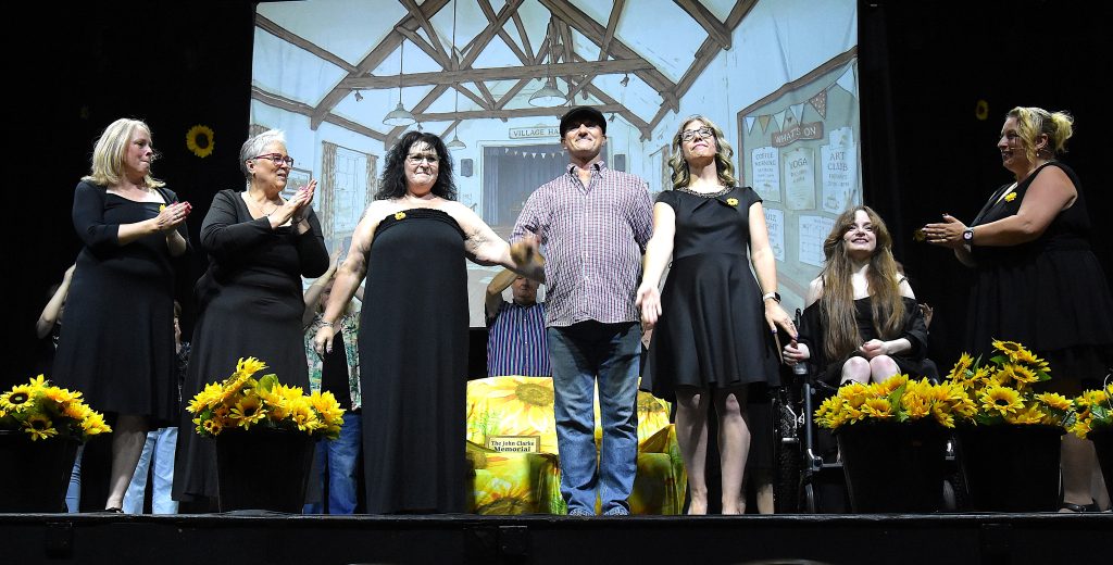 The principal cast of Calendar Girls: The Musical take a bow during the curtain call at The Carnegie, Thetford. The performers stand hand in hand at the front of the stage, applauded by fellow cast members behind them, with sunflower displays lining the stage and the village hall set projected in the background.