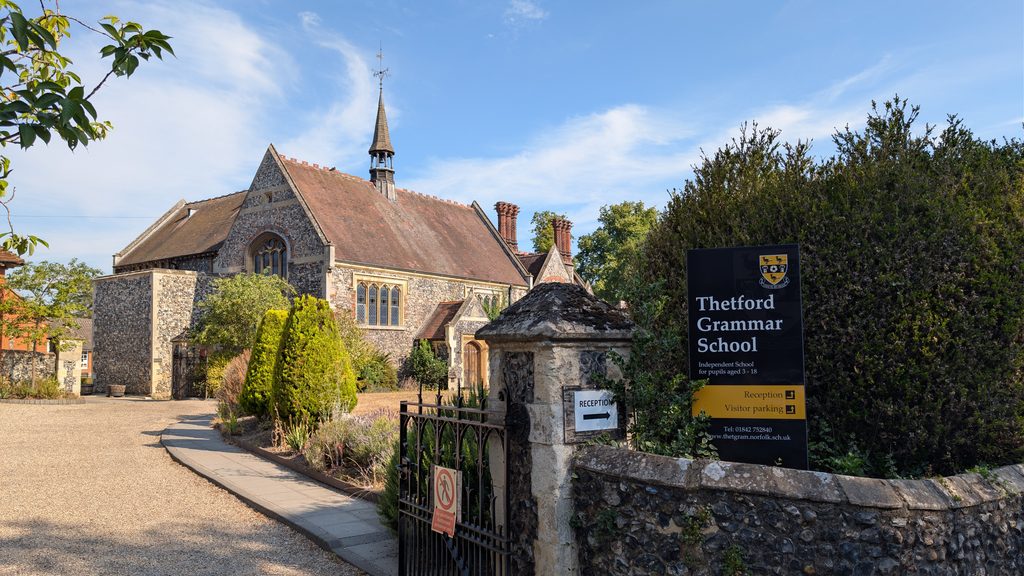 Exterior of Thetford Grammar School on a sunny day, showing the historic flint school building with its steep tiled roof and small spire, landscaped grounds and the school entrance sign in the foreground.
