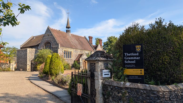Exterior of Thetford Grammar School on a sunny day, showing the historic flint school building with its steep tiled roof and small spire, landscaped grounds and the school entrance sign in the foreground.