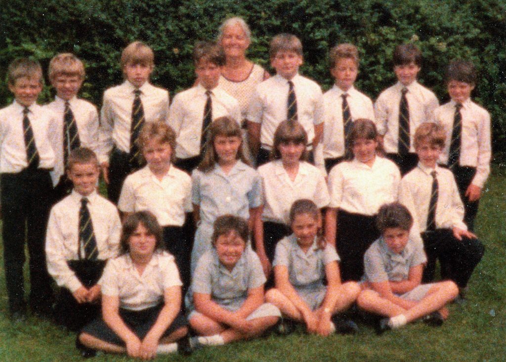 Colour school photograph of a mixed group of pupils and a teacher posed outdoors on grass. Pupils wear school uniform, with boys in white shirts and striped ties and girls in white or pale blue blouses.